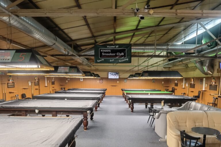 Ein Snooker-Club mit mehreren überdachten Snooker-Tischen, einem grün beleuchteten Tisch im hinteren Bereich, Sofas und einem Schild mit der Aufschrift Home of KENNYS Snooker Club. An der Decke sind warme Beleuchtung und freiliegende Rohrleitungen zu sehen.