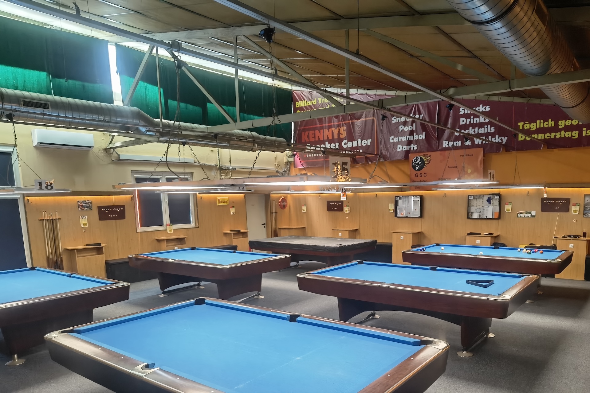 Eine geräumige Billardhalle mit mehreren Billardtischen aus blauem Filz, Deckenbeleuchtung und holzgetäfelten Wänden. Bunte Schilder werben für Spiele, Snacks und Getränke. Das Sonnenlicht fällt durch die grünen Fensterläden.
