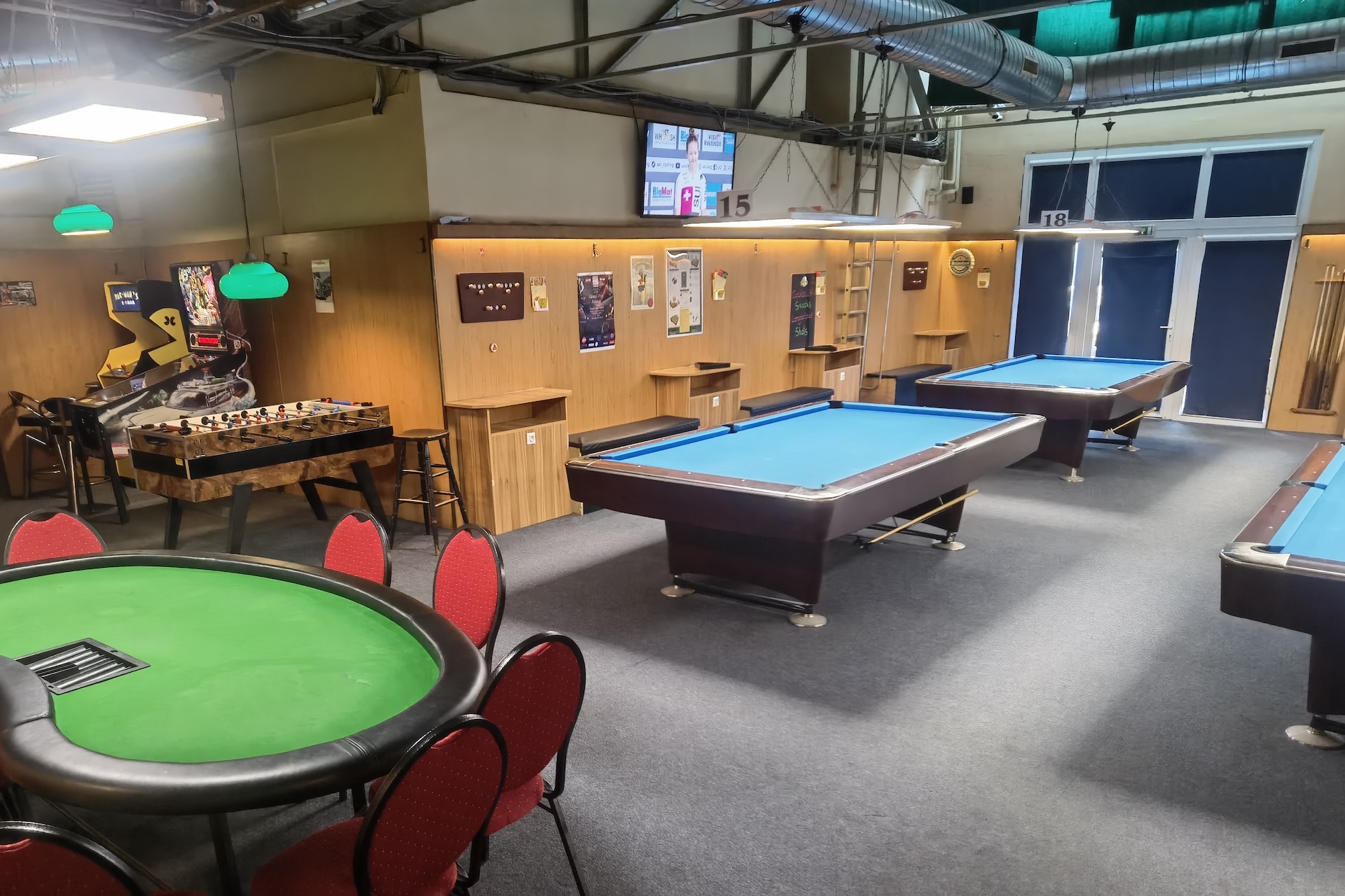 Ein Freizeitraum mit Billardtischen, einem Kickertisch, einem Pokertisch mit roten Stühlen, Spielautomaten und Postern an holzgetäfelten Wänden unter freiliegenden industriellen Deckenrohren.