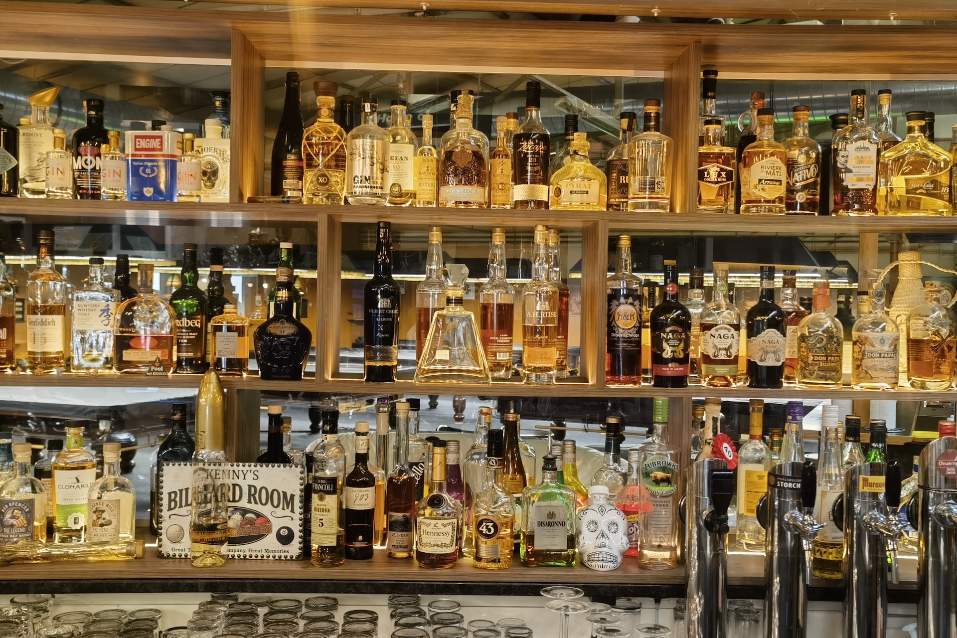 Regale mit einer Vielzahl von Spirituosenflaschen, darunter Whiskey, Tequila, Rum und Wodka, hinter einer Bar mit Gläsern und Getränkeautomaten im Vordergrund.