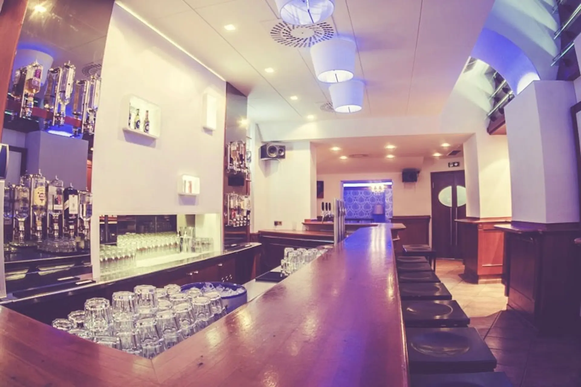 Eine moderne Bar mit einem langen Holztresen, Barhockern, Regalen mit Flaschen und ordentlich gestapelten Gläsern. Die warme und blaue Beleuchtung schafft eine entspannte, einladende Atmosphäre.
