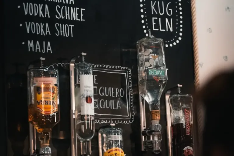 Mehrere umgedrehte Spirituosenflaschen sind auf einem Spender vor einer schwarzen Kreidetafel mit handgeschriebenen Getränkeoptionen in deutscher Sprache montiert, darunter Wodka Schnee Shot und Tequila.