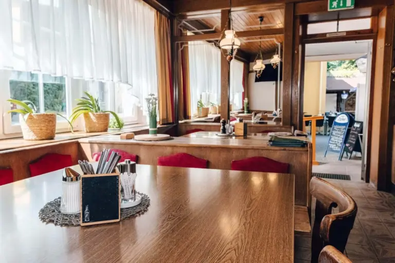 kirchenwirt jöch Ein gemütliches Restaurant mit Holztischen und -stühlen, roten Sitzpolstern, Topfpflanzen auf den Fensterbänken und Sonnenlicht, das durch große Fenster mit weißen Vorhängen fällt. Vor der Tür ist ein Schild mit der Speisekarte zu sehen.