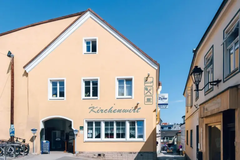 Gelbes Gebäude mit der Aufschrift Kirchenwirt an der Fassade, mit weiß gerahmten Fenstern und einem kleinen Bogeneingang. Es liegt an einer schmalen Straße mit klarem, blauem Himmel über sich.