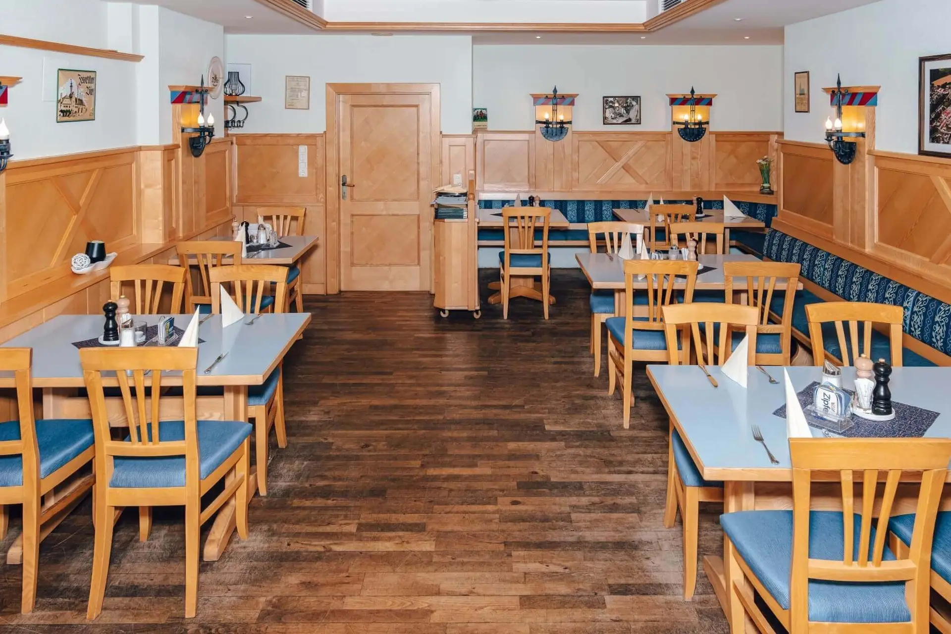 Ein kleines, leeres Restaurant mit Holztischen und -stühlen, blauen Sitzkissen, ordentlich gedeckten Tischen mit Servietten und hölzerner Wandverkleidung. Gemälde und Wandlampen schmücken die hellen Wände.