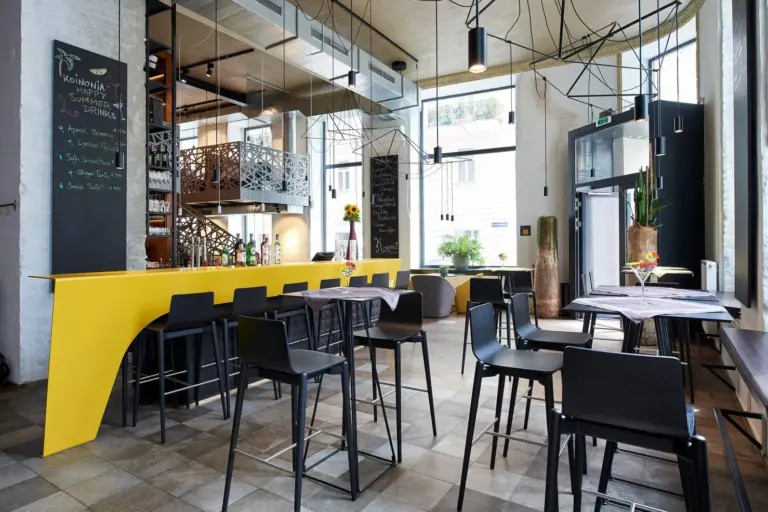 Modernes Café mit hohen Decken, großen Fenstern, schwarzen Stühlen, einem gelben Bartresen, Hängelampen und Kreidetafeln an den Wänden. Natürliches Licht durchflutet den stilvollen, im industriellen Stil eingerichteten Raum.