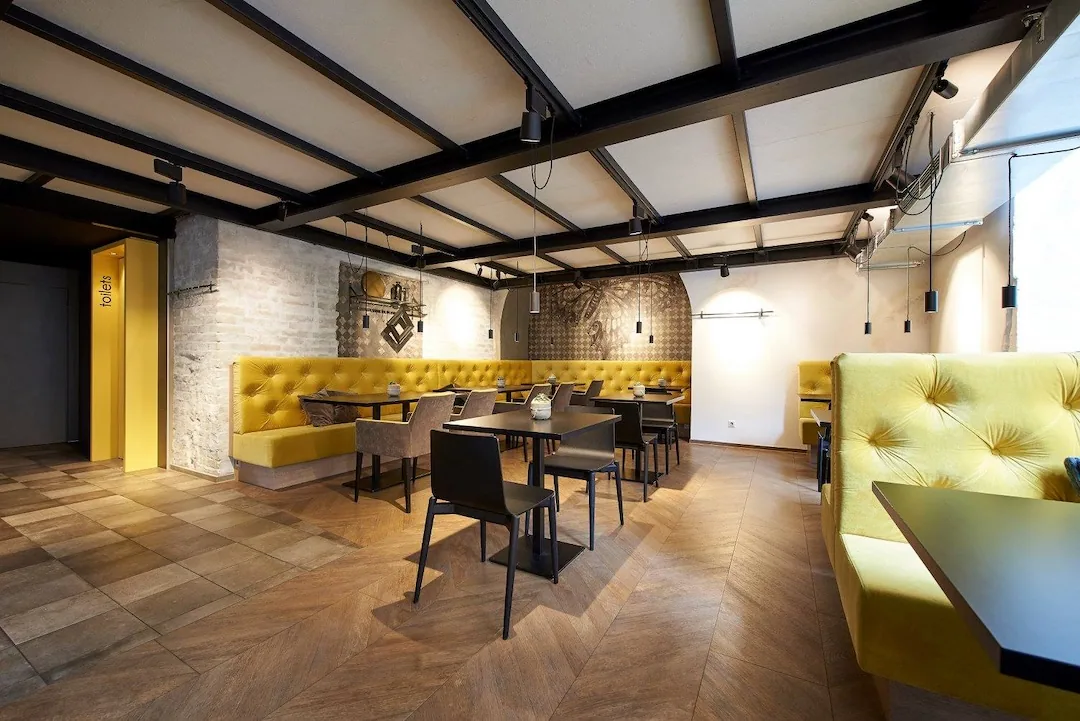 Modernes Restaurantinterieur mit gelben getufteten Sitzbänken, schwarzen Tischen und Stühlen, hellem Holzfußboden im Fischgrätmuster, freiliegender Ziegelwand und industrieller Decke. Der Raum erscheint hell und sauber mit minimalistischem Dekor.