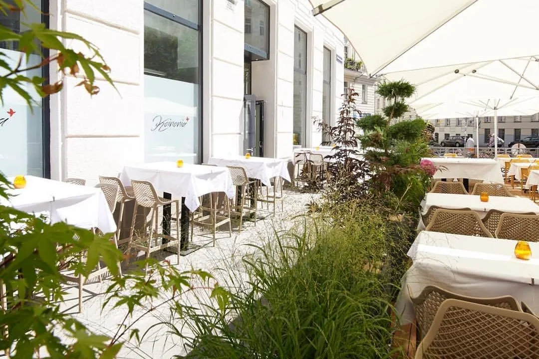Eine helle Restaurantterrasse mit weißen Tischen und Korbstühlen, beschattet von großen Sonnenschirmen und umgeben von Grünpflanzen am Rande des Sitzbereichs.