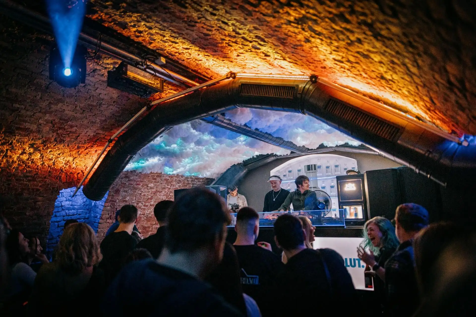 Ein Publikum genießt die Musik in einem schwach beleuchteten Underground-Club mit Backsteinwänden und gewölbter Decke, während zwei DJs in einer Kabine bei dramatischer Beleuchtung auflegen.