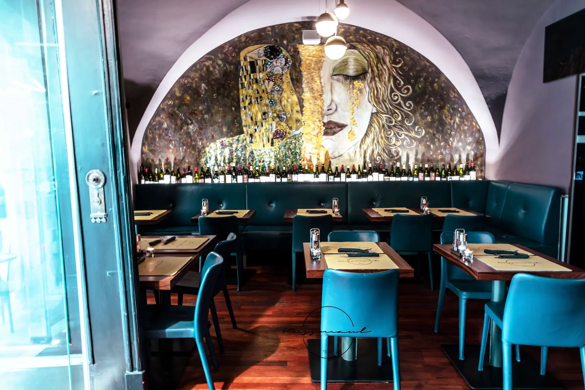 Ein modernes Restaurant mit tealfarbenen Stühlen und Holztischen, die zum Essen einladen. An der Rückwand befindet sich ein großes, von Gustav Klimt inspiriertes Wandgemälde, unter dem viele Weinflaschen aufgereiht sind. Die sanfte Beleuchtung schafft eine gemütliche Atmosphäre.