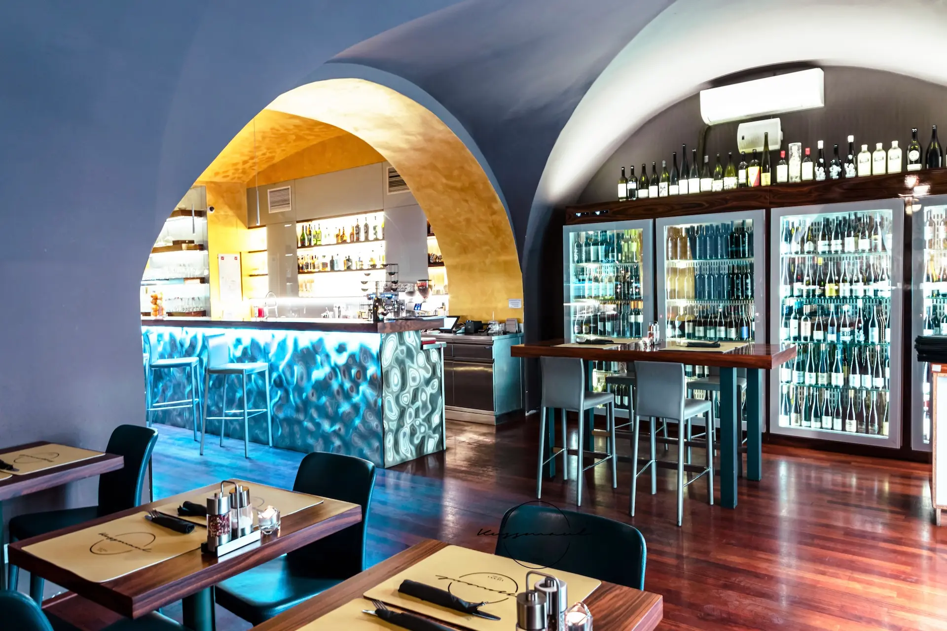 Modernes Restaurantinterieur mit gewölbten Decken, Holzböden und einer blau leuchtenden Bar. Die Weinflaschen stehen in Glasvitrinen, und die Tische sind mit Tischsets und Gewürzen gedeckt.