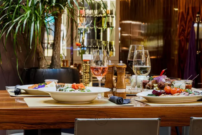 Ein hölzerner Restauranttisch mit Tellern mit Gourmetgerichten, zwei Gläsern mit Weißwein, Salz- und Pfeffermühlen und einer Topfpflanze im Hintergrund. Die Szene suggeriert ein feines Essen in einem eleganten Rahmen.