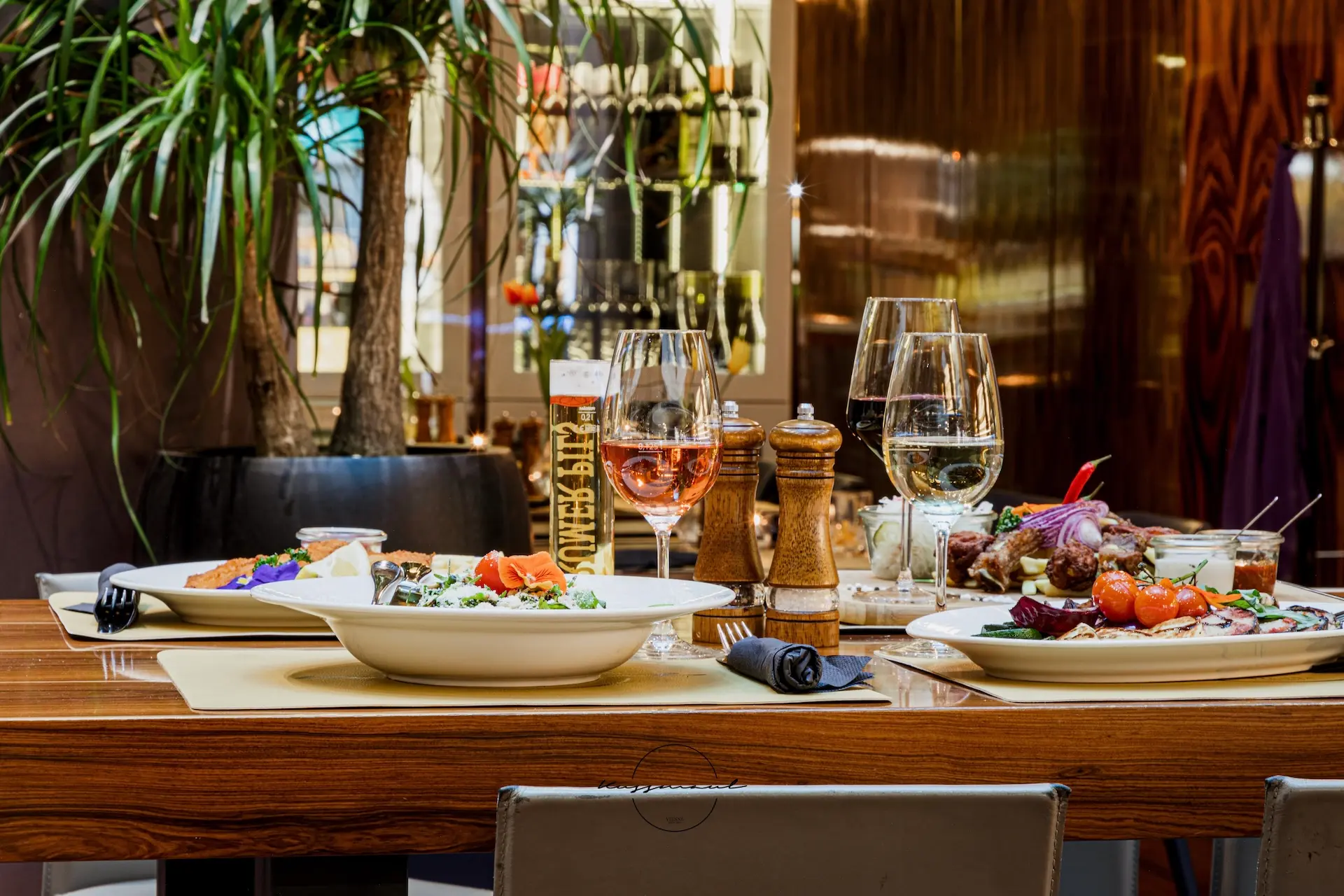 Ein hölzerner Restauranttisch mit Tellern mit Gourmetgerichten, zwei Gläsern mit Weißwein, Salz- und Pfeffermühlen und einer Topfpflanze im Hintergrund. Die Szene suggeriert ein feines Essen in einem eleganten Rahmen.