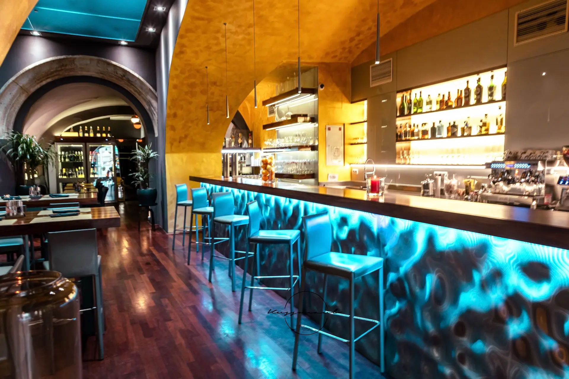 Modernes Barinterieur mit blau beleuchtetem Bartresen, hohen Metallhockern, Holzboden, gewölbter Decke, Schnapsflaschen in Regalen und einer stilvollen, gemütlichen Atmosphäre.