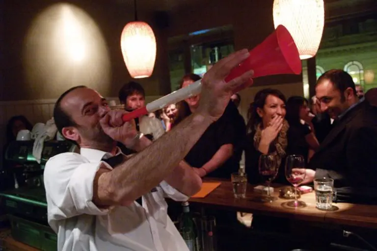 Ein Mann in einem weißen Hemd bläst begeistert in eine rot-weiße Vuvuzela an einer Bar, während eine Gruppe von Menschen hinter dem Tresen lacht und mit Getränken in der Hand unter warmen Hängelampen zusieht.