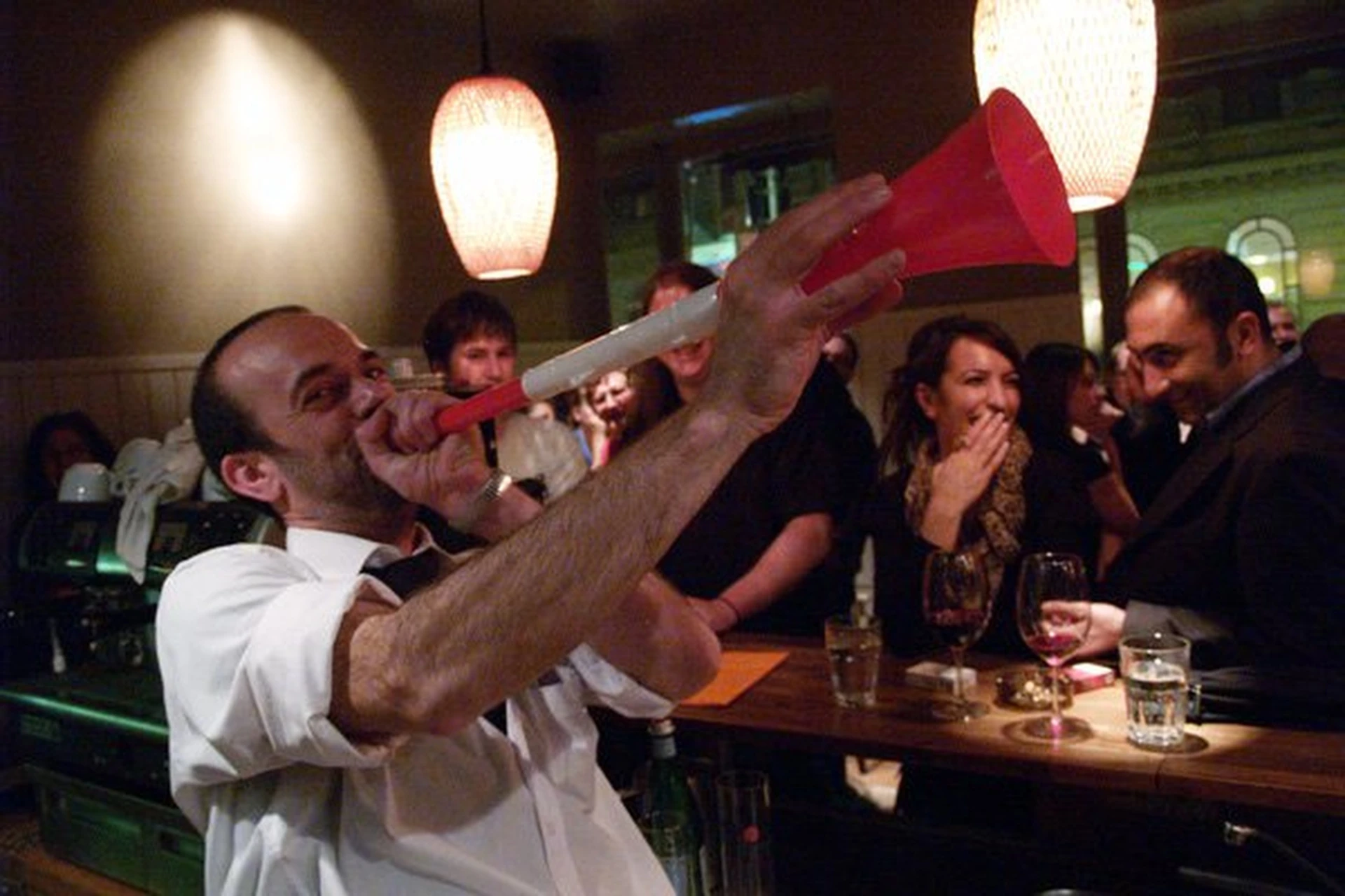 Ein Mann in einem weißen Hemd bläst begeistert in eine rot-weiße Vuvuzela an einer Bar, während eine Gruppe von Menschen hinter dem Tresen lacht und mit Getränken in der Hand unter warmen Hängelampen zusieht.