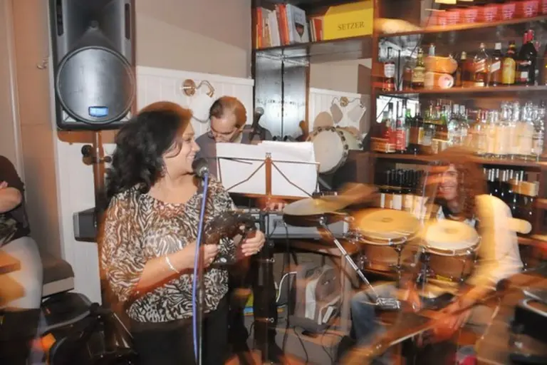 Eine Frau singt in ein Mikrofon, während sie ein Tamburin in der Hand hält, begleitet von Musikern, die Keyboard und Schlagzeug spielen, in einer gemütlichen, warm beleuchteten Bar oder einem Café, das mit Regalen voller Flaschen und Bücher gefüllt ist.