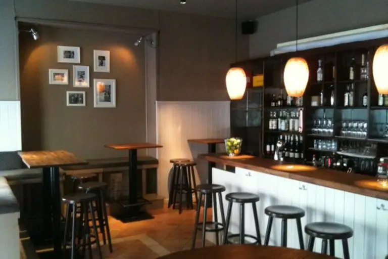 Eine gemütliche Bar mit Holztischen und -hockern, warmer Beleuchtung durch Hängelampen, Regalen mit Flaschen und Gläsern und gerahmten Fotos an der Wand. Die Atmosphäre ist sauber und einladend.