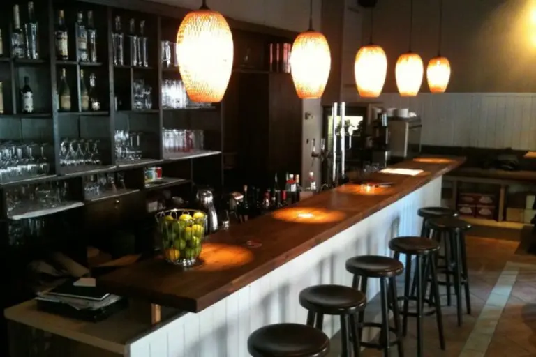 Eine gemütliche Bar mit einer hölzernen Arbeitsplatte, fünf Barhockern, darüber hängenden Hängelampen und Regalen mit Gläsern und Flaschen im Hintergrund. Eine Schale mit grünen Äpfeln steht auf dem Tresen.