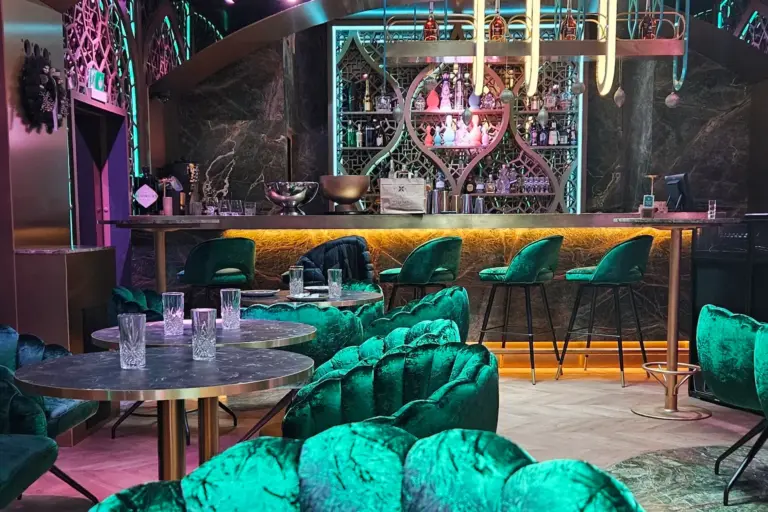 Eine stilvolle, moderne Bar mit grünen Samtstühlen, runden Marmortischen, Neonbeleuchtung und dekorativen Regalen mit Flaschen hinter dem Bartresen. Mehrere leere Gläser stehen auf den Tischen und der Bar.