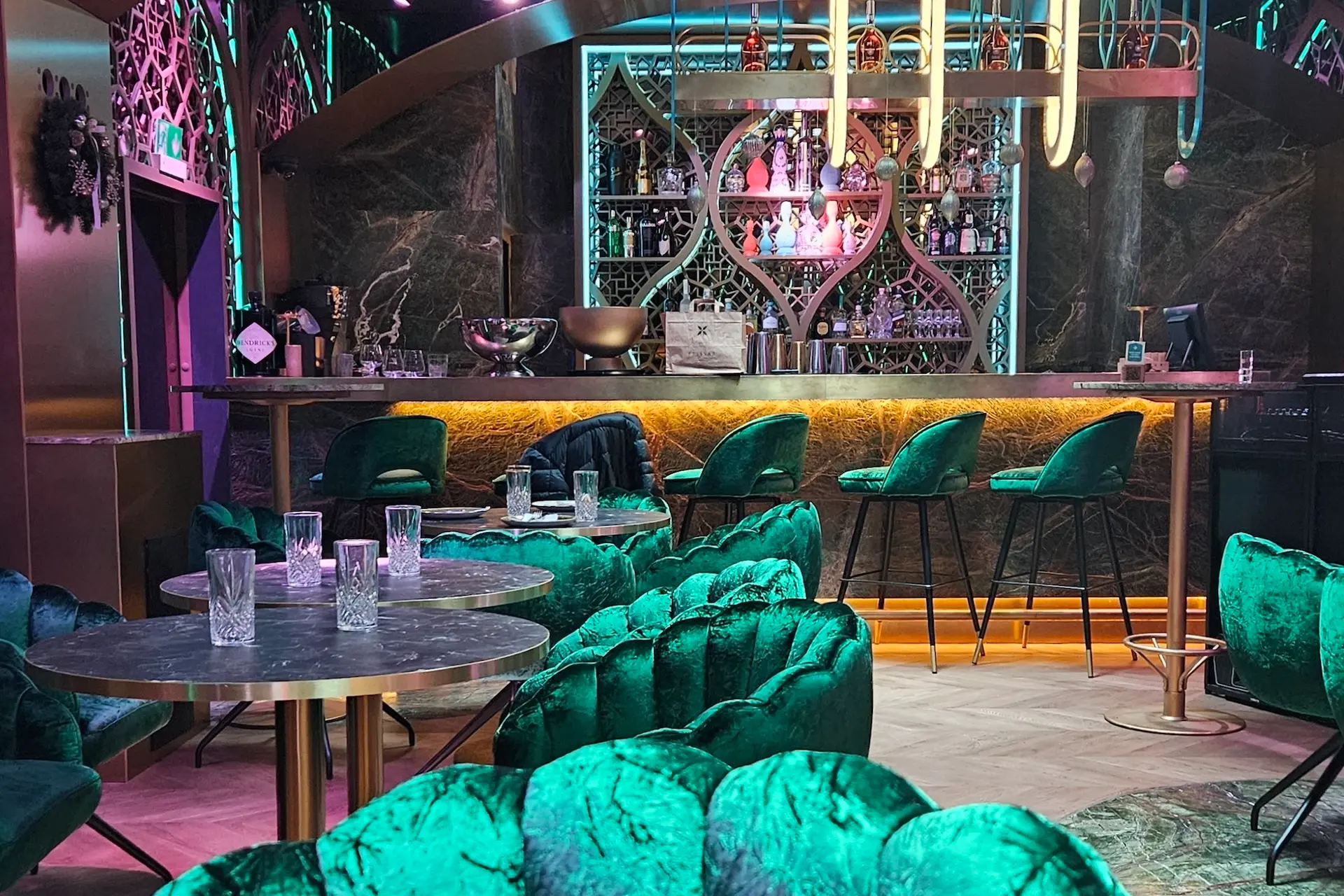 Eine stilvolle, moderne Bar mit grünen Samtstühlen, runden Marmortischen, Neonbeleuchtung und dekorativen Regalen mit Flaschen hinter dem Bartresen. Mehrere leere Gläser stehen auf den Tischen und der Bar.