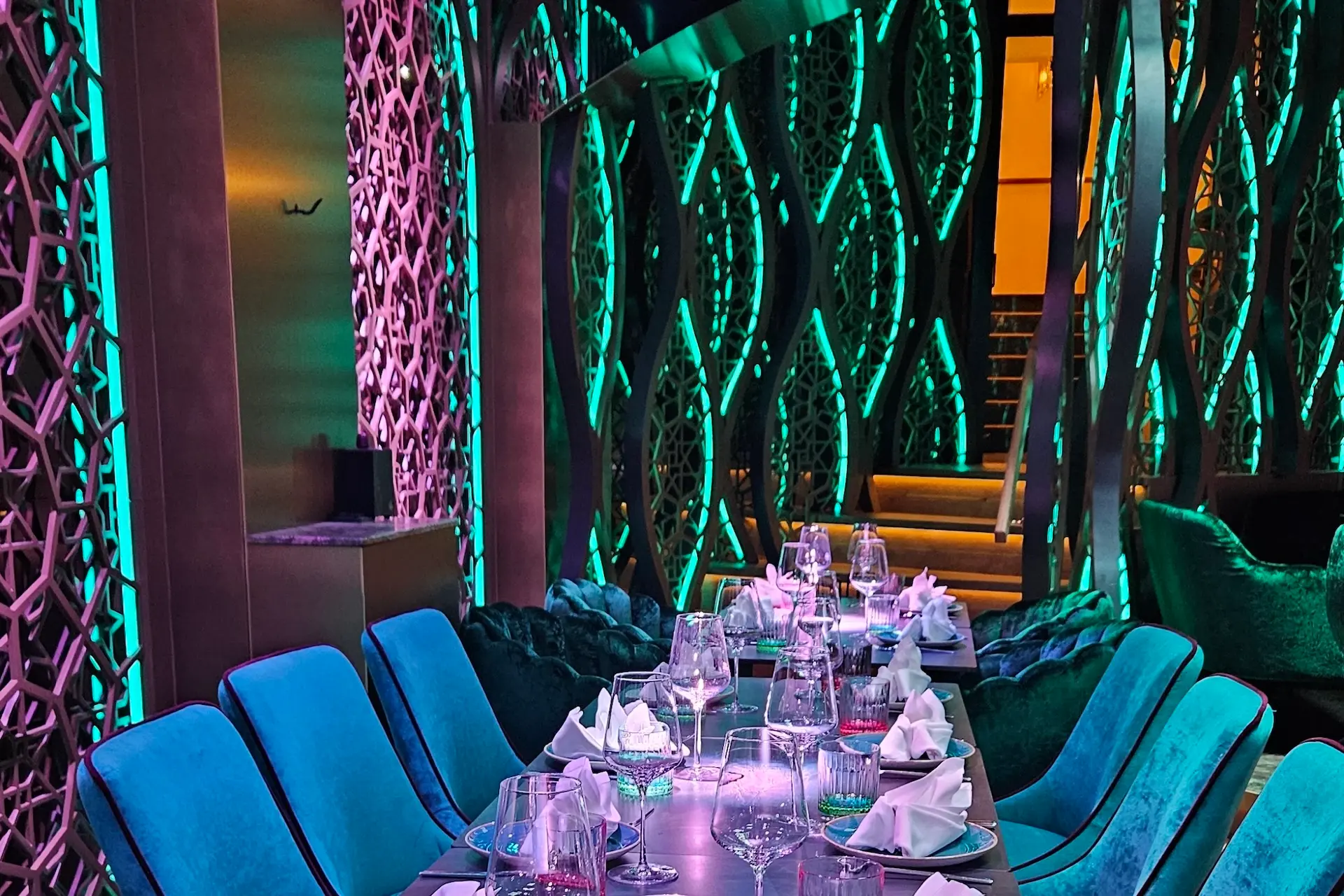 Ein moderner Restaurantbereich mit einem langen Tisch für die Gäste, blauen Stühlen, gefalteten Servietten, Weingläsern und leuchtend blaugrüner Neonbeleuchtung mit dekorativen Mustern an den Wänden.