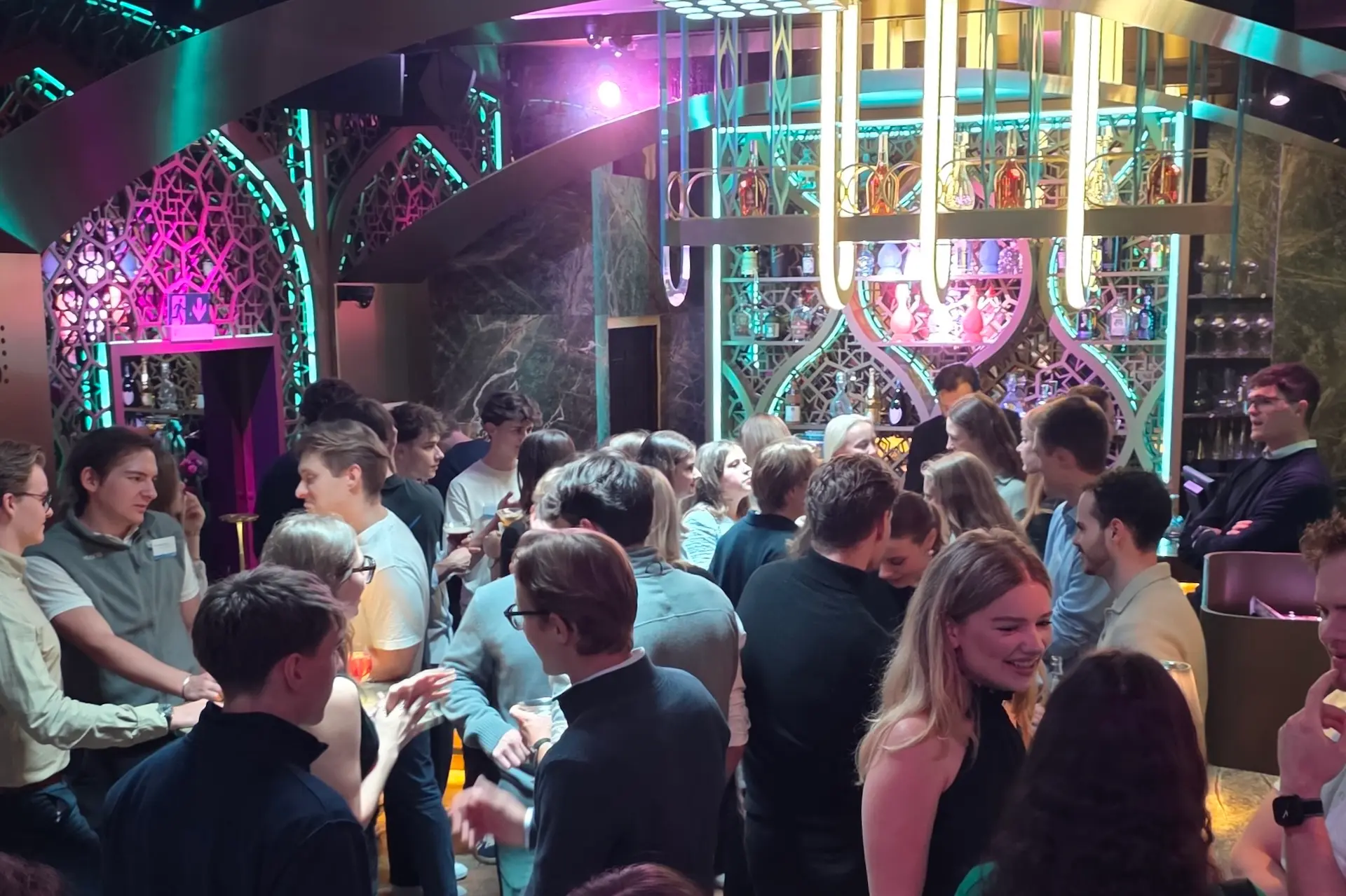 Eine lebhafte Gruppe junger Erwachsener trifft sich in einer lebhaften, modernen Bar mit bunter Beleuchtung und dekorativen Tafeln. Einige Leute unterhalten sich in Gruppen, während andere an der stylischen Bar stehen.