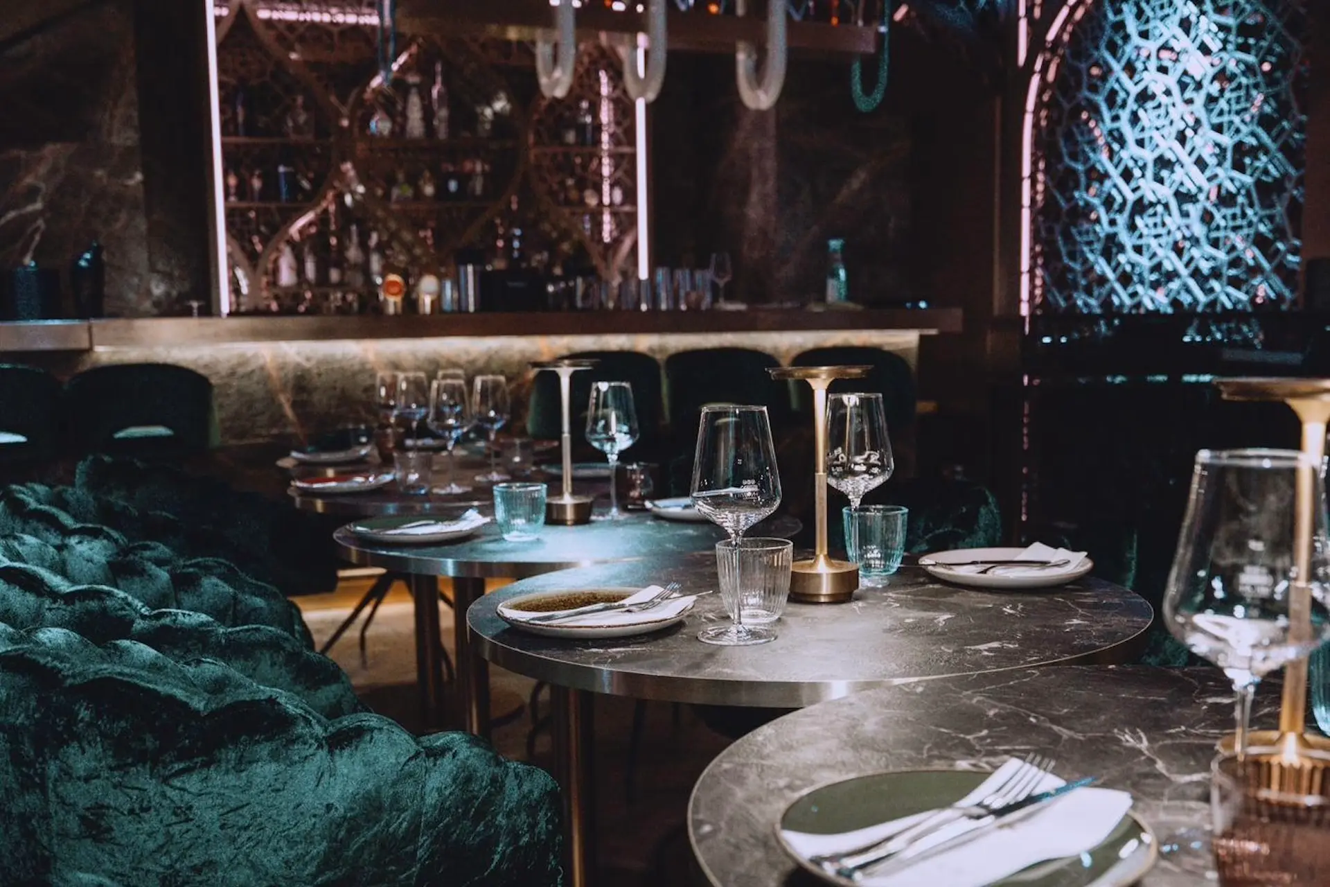 Ein stilvolles, schummriges Restaurant mit Marmortischen, die mit Tellern, Gläsern und Besteck gedeckt sind. Plüschige grüne Samtstühle umgeben die Tische, und im Hintergrund ist eine mit Flaschen bestückte Bar zu sehen.