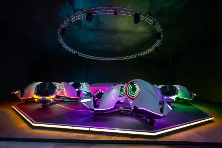 Eine Gruppe futuristischer, neonbeleuchteter Arcade-Fahrzeuge, die schnittigen Motorrädern ähneln, sind in einem dunklen Raum unter einem runden Metallgestell angeordnet und leuchten in leuchtenden Farben wie Lila, Grün, Gelb und Pink.