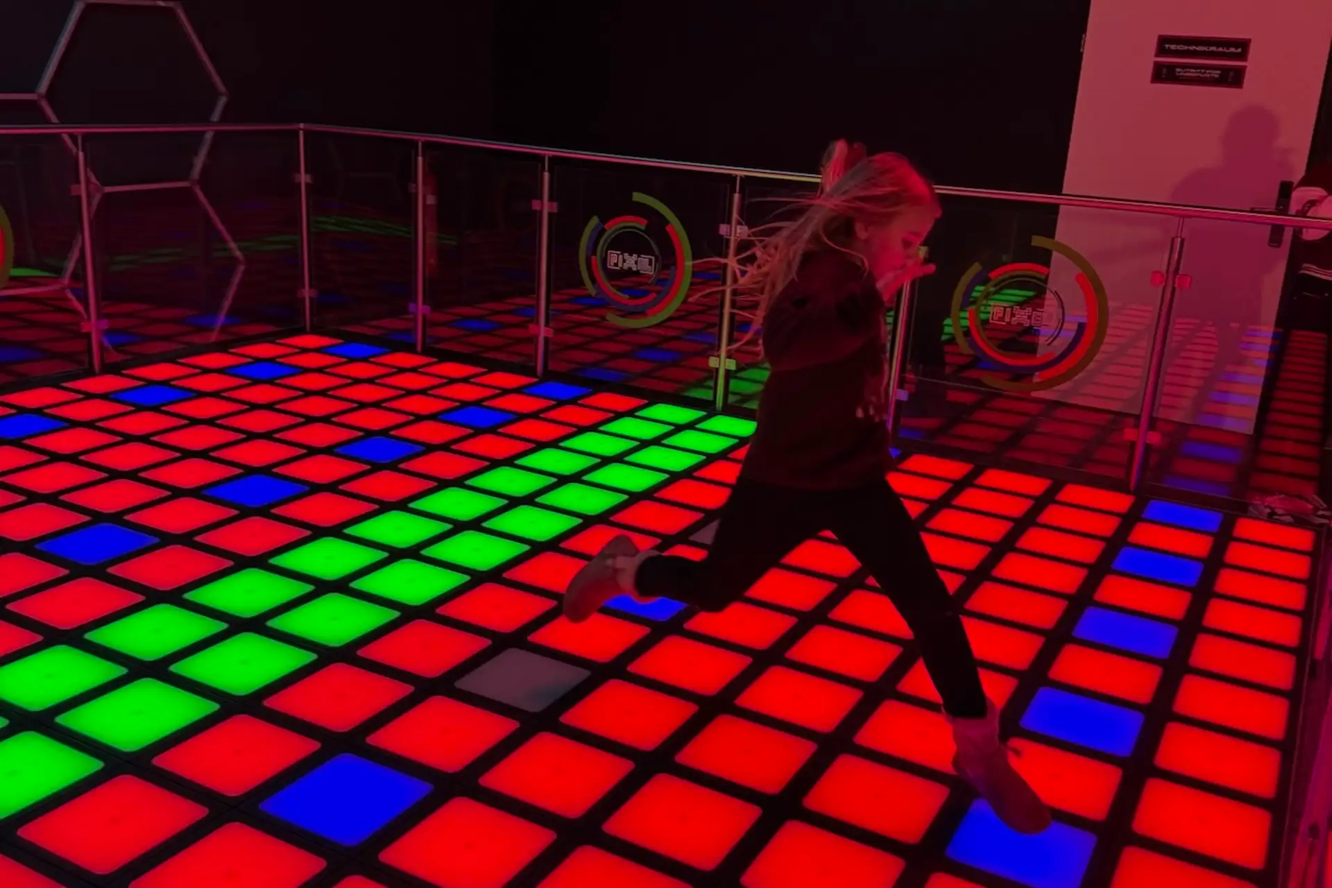 Ein Kind mit langen Haaren springt über einen hell erleuchteten Boden mit großen roten, grünen und blauen Quadraten in einem Indoor-Spielbereich. Im Hintergrund sind Glasbarrieren und bunte Schilder an der Wand zu sehen.