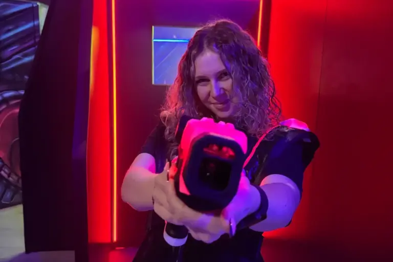 Eine Person mit lockigem Haar lächelt, während sie in einem dunklen Raum mit leuchtend roten und violetten Neonröhren eine Laser-Tag-Pistole ausrichtet.