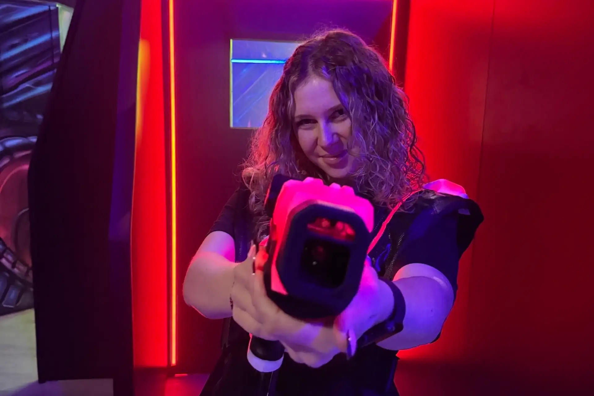 Eine Person mit lockigem Haar lächelt, während sie in einem dunklen Raum mit leuchtend roten und violetten Neonröhren eine Laser-Tag-Pistole ausrichtet.
