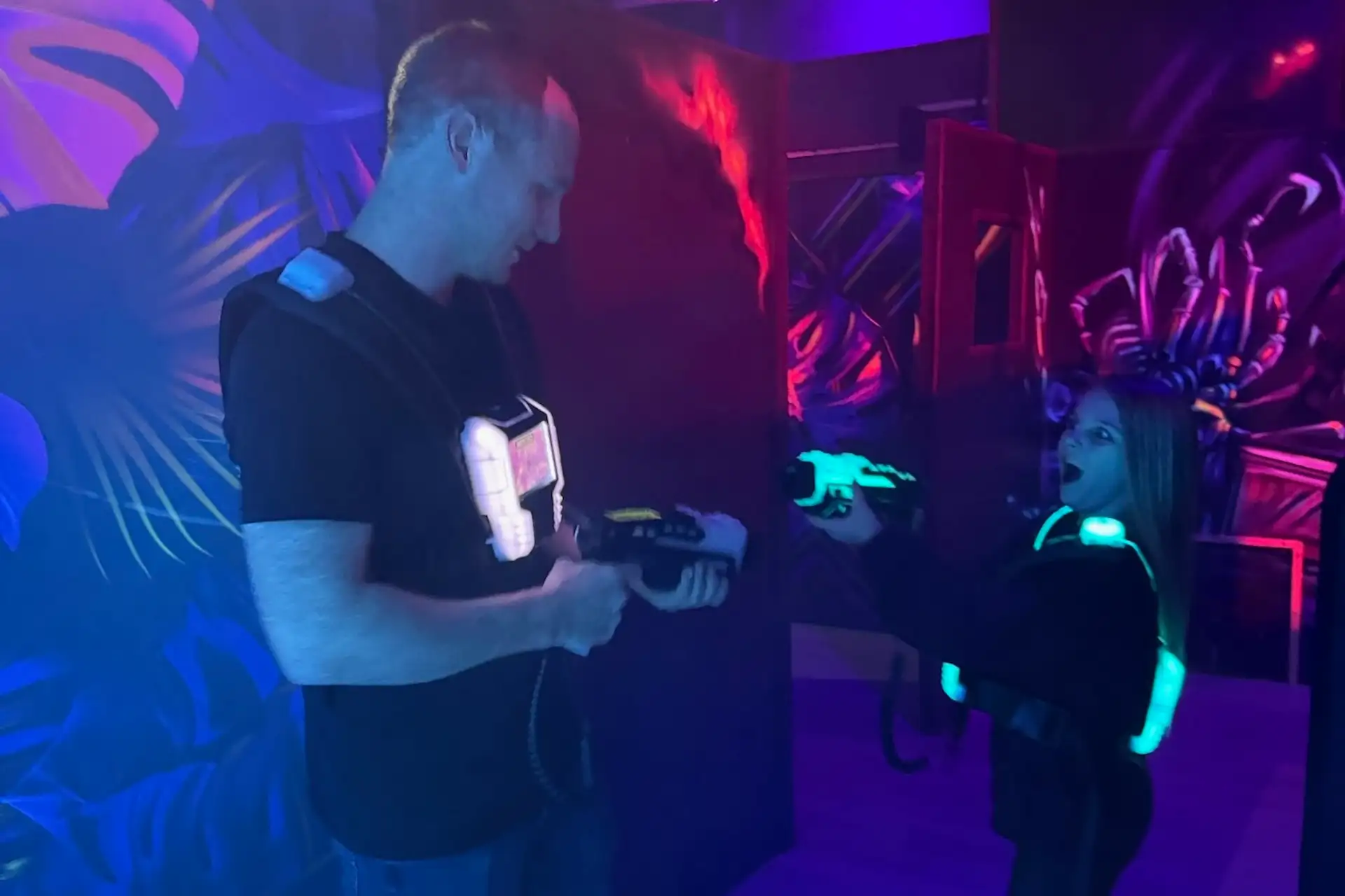 Ein Erwachsener und ein Kind, die Laser-Tag-Westen tragen und Laserpistolen in der Hand halten, stehen sich in einer dunklen, neonbeleuchteten Arena mit leuchtenden, bunten Wänden gegenüber.