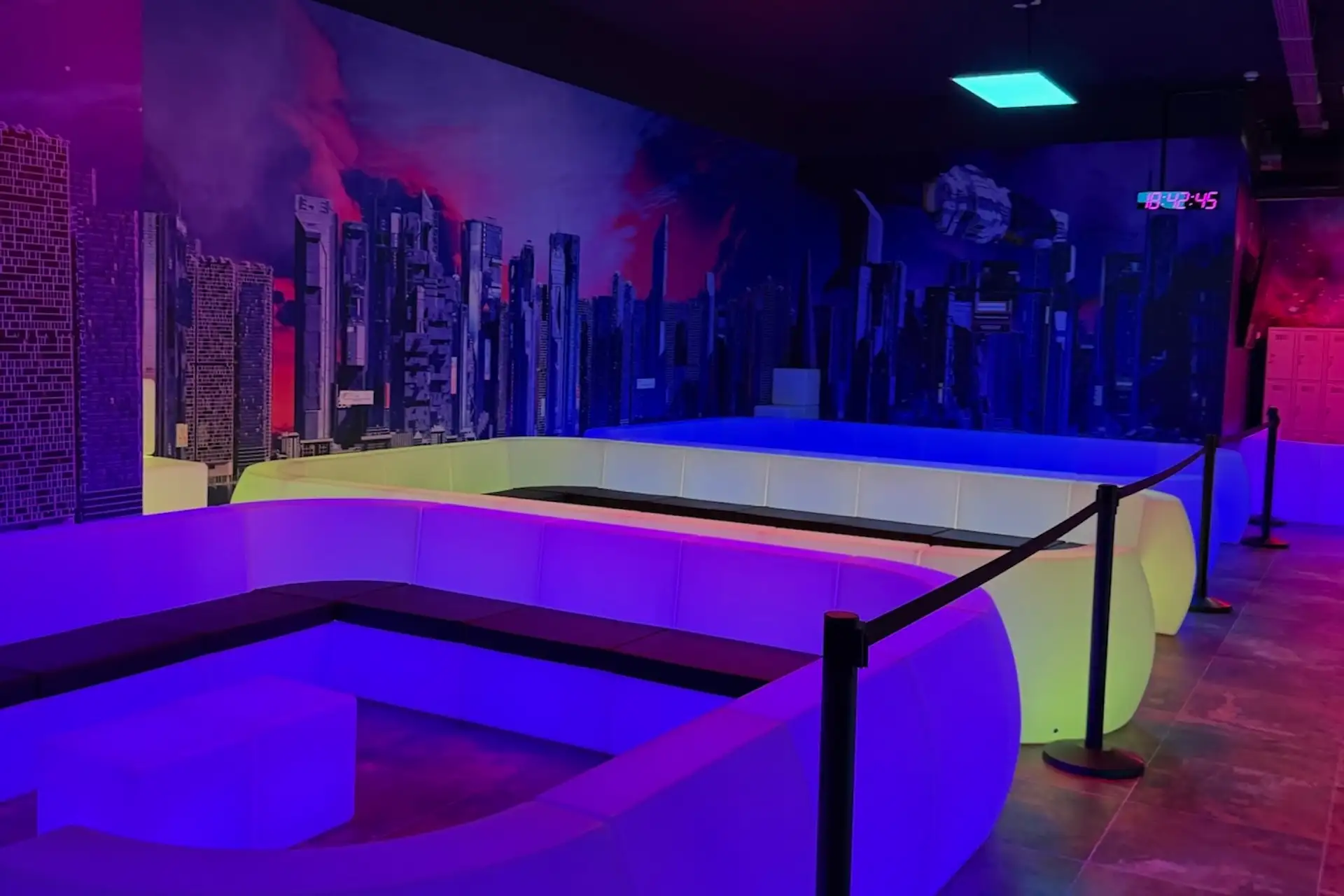 Eine moderne Lounge mit geschwungenen, weißen Sitzmöbeln, die von bunten Neonröhren beleuchtet werden. An den Wänden ist ein futuristisches Stadtbild zu sehen, und Samtseile trennen den Sitzbereich vom Rest des Raums.