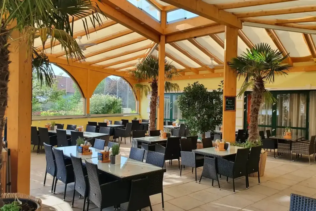 Eine geräumige, überdachte Restaurantterrasse mit Tischen mit Glasplatte, schwarzen Korbstühlen, Topfpflanzen und Palmen unter einer hölzernen Pergola. Das Sonnenlicht strömt durch große Fenster und ein offenes Dach herein.