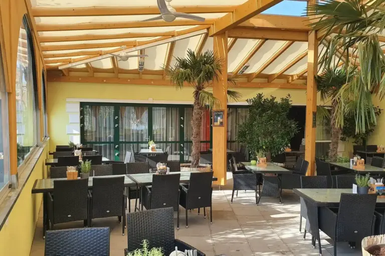 Eine überdachte Außenterrasse in einem Restaurant mit schwarzen Korbtischen und -stühlen, Topfpflanzen und großen Fenstern. Das Sonnenlicht fällt durch das hölzerne Pergoladach und schafft eine helle und einladende Atmosphäre.