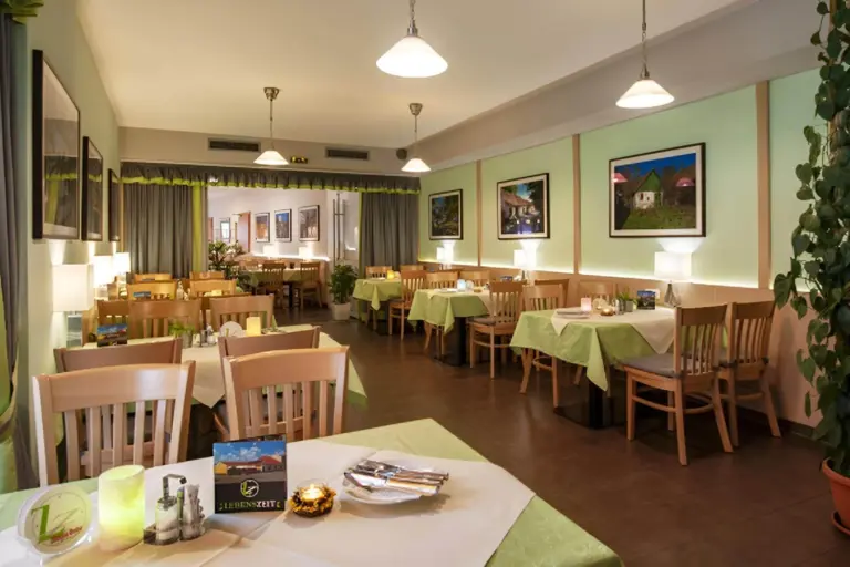 Ein gemütlicher Restaurantbereich mit Holztischen und -stühlen, grün-weißen Tischtüchern, gerahmten Fotos an pastellgrünen Wänden, Hängelampen und Topfpflanzen, die eine einladende Atmosphäre schaffen.