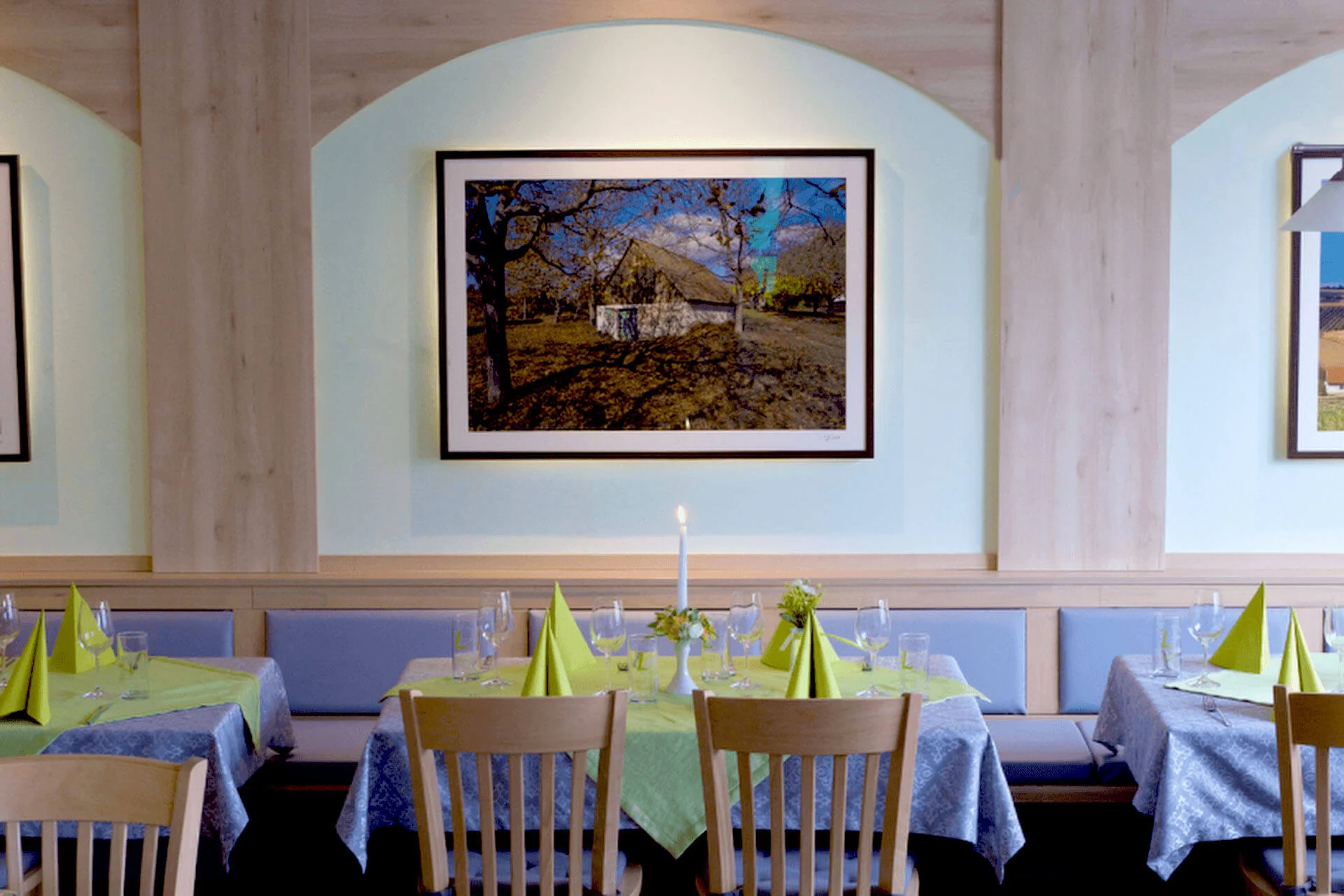 Ein Restaurant mit Holzstühlen und Tischen, die mit grünen Servietten, Tischtüchern, Gläsern und einer einzigen Kerze gedeckt sind. Ein gerahmtes Landschaftsfoto hängt an einer pastellfarbenen Wand.