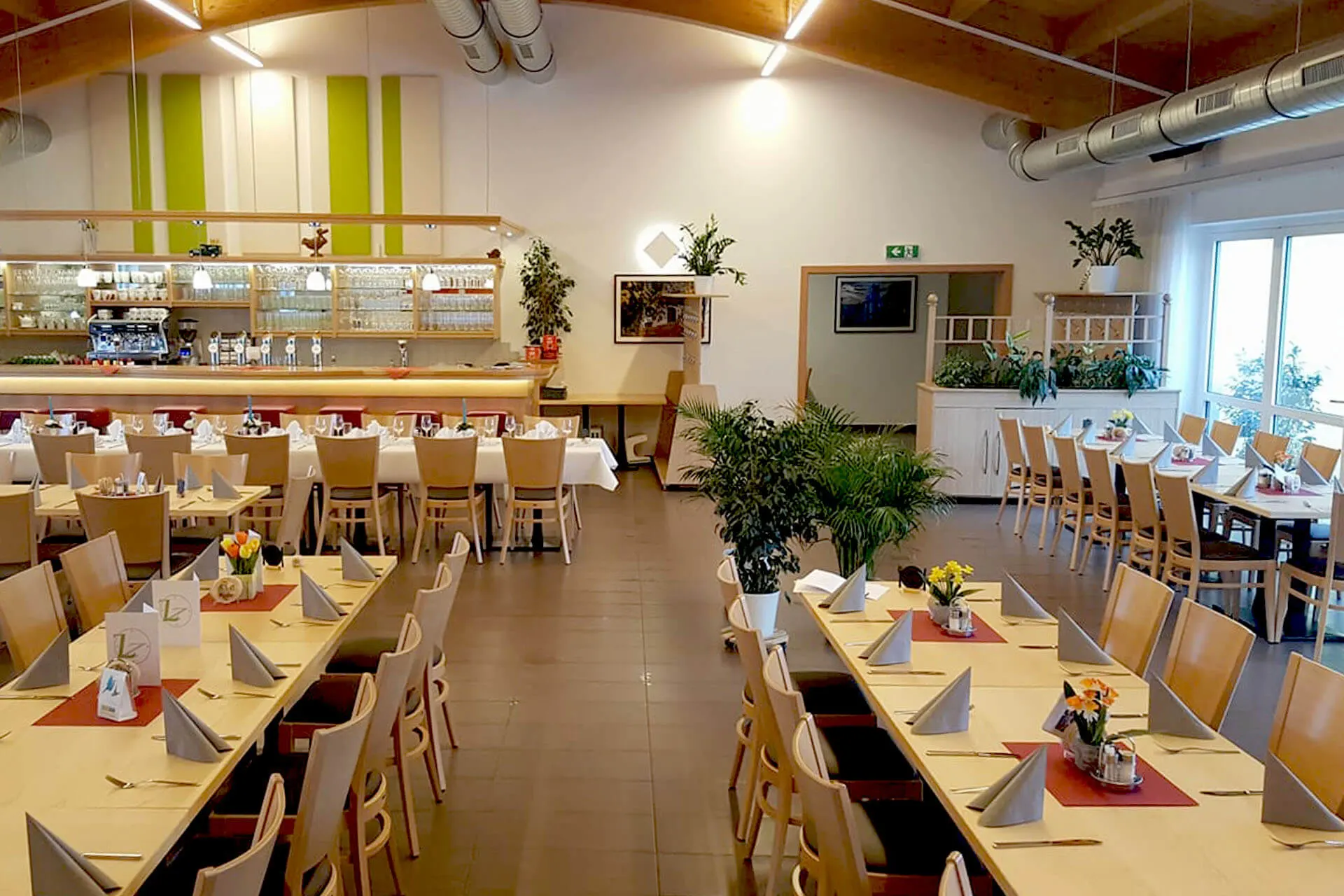 Geräumiger, moderner Speisesaal mit Holzstühlen und Tischen, die mit Servietten, Speisekarten und kleinen Blumenvasen gedeckt sind. Helle Beleuchtung, Grünpflanzen, ein Barbereich und große Fenster sorgen für eine einladende Atmosphäre.