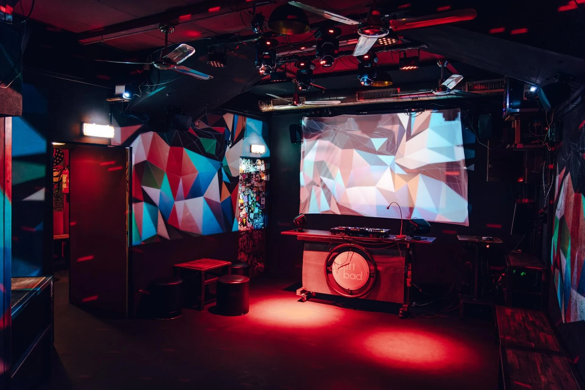 Luftbad Ein schwach beleuchteter Nachtclub mit roter Beleuchtung, einer DJ-Kabine, geometrisch gemusterter Wandkunst, leeren Hockern und einem großen Bildschirm, auf dem bunte abstrakte Formen zu sehen sind.