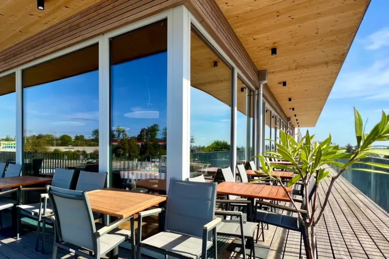 Essbereich im Freien mit Holztischen und -stühlen auf einer Terrasse neben einem modernen Gebäude mit großen Glasfenstern und einem schrägen Holzdach, unter einem klaren blauen Himmel. Eine Topfpflanze steht auf der Terrasse.