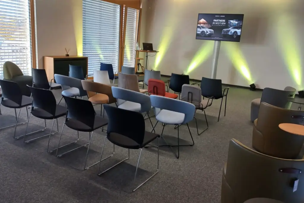 Ein moderner Konferenzraum mit leeren Stuhlreihen vor einem großen Bildschirm, auf dem Autos zu sehen sind, und einem Laptop auf einem Podium; gelbe Strahler beleuchten die Wände und Jalousien lassen natürliches Licht herein.