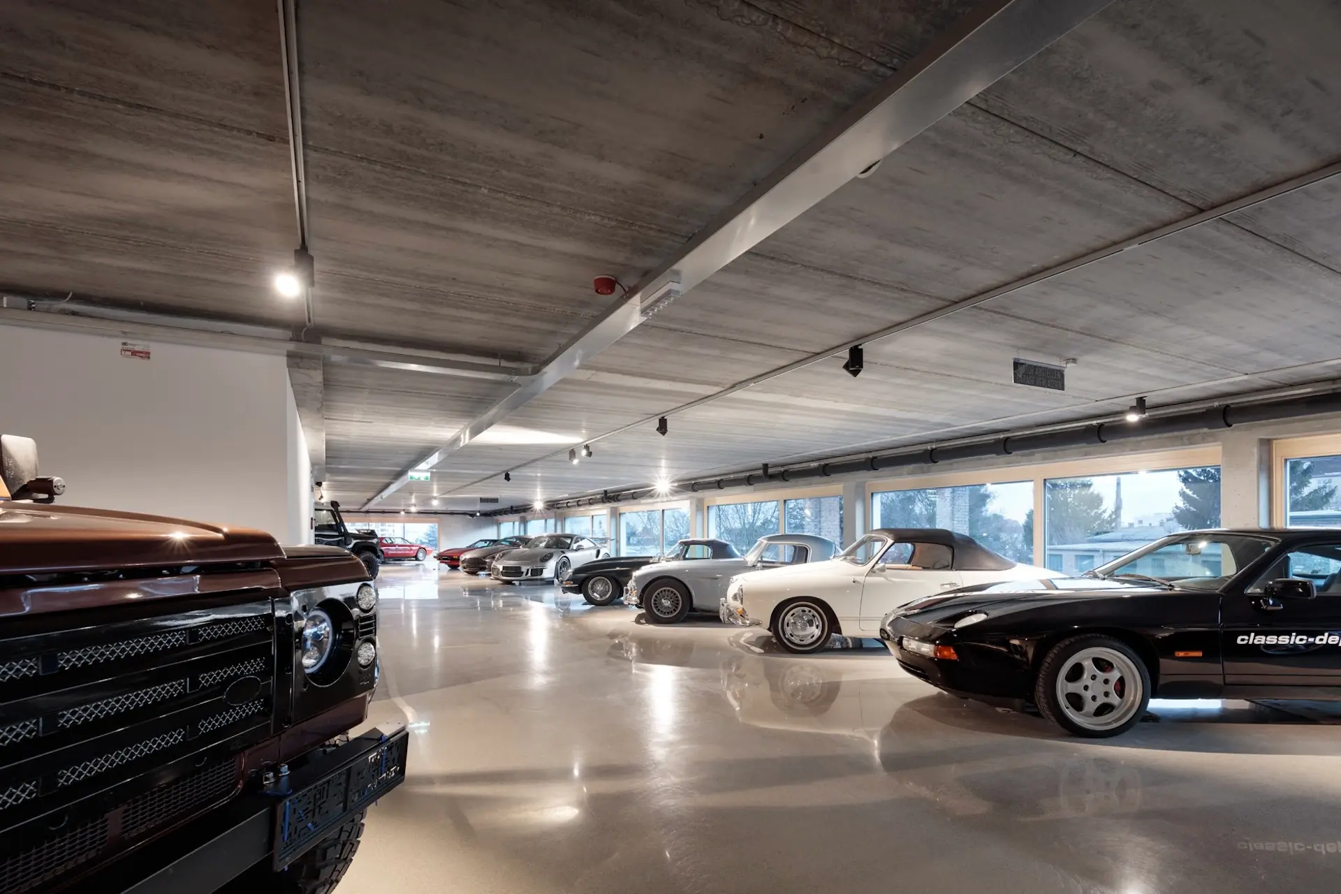 Eine moderne, gut beleuchtete Garage mit polierten Betonböden und großen Fenstern, in der eine Reihe von Oldtimern und Klassikern in verschiedenen Farben, darunter Schwarz, Weiß und Silber, ausgestellt sind.