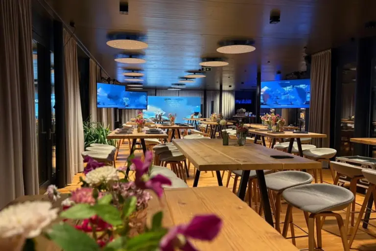 Ein moderner Konferenzraum mit Holztischen, gepolsterten Hockern und Blumenkränzen in der Mitte. Große Bildschirme zeigen ein blaues Bild an der Vorderseite, und der Raum ist mit runden Deckenleuchten sanft beleuchtet.