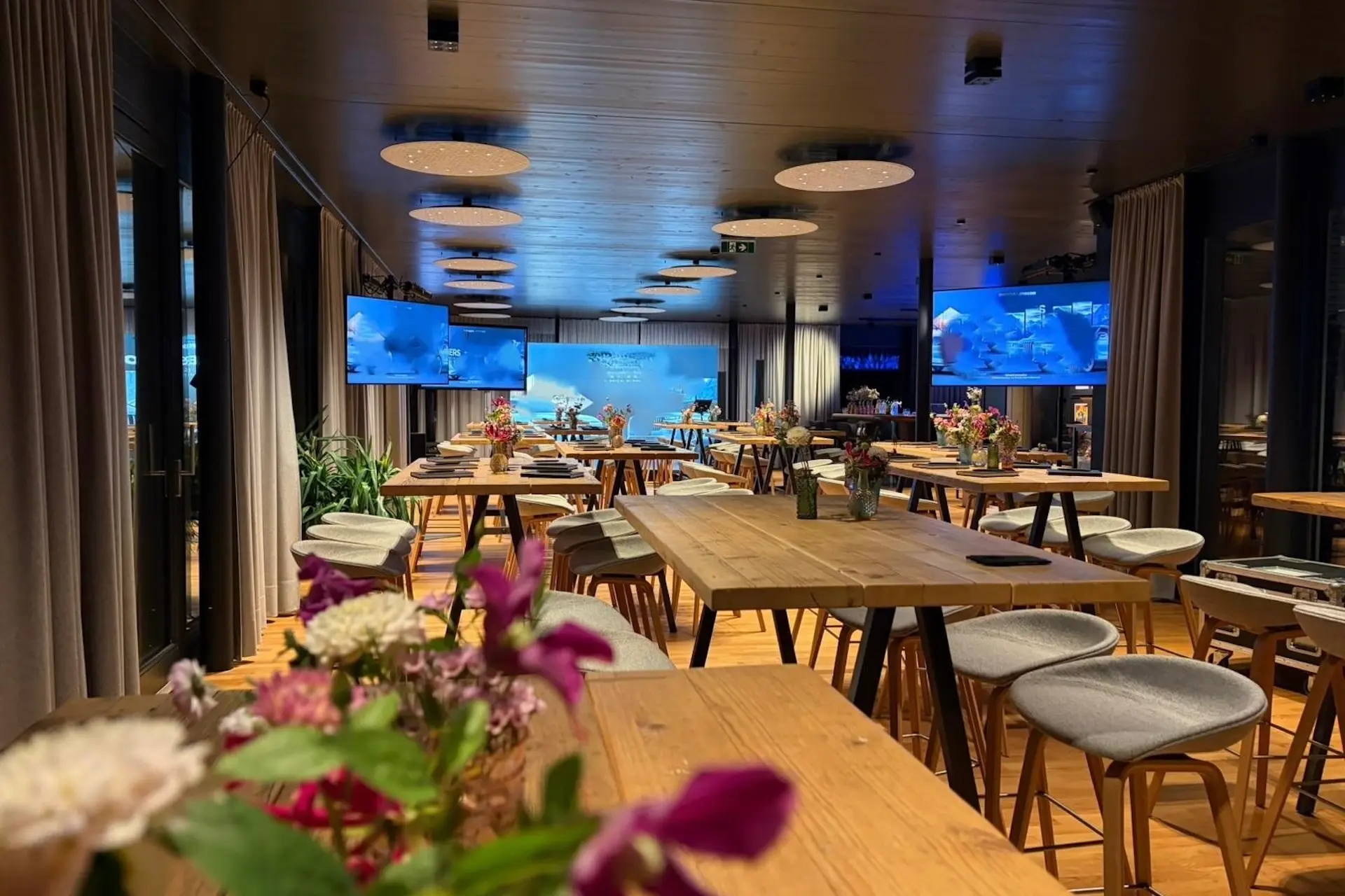 Ein moderner Konferenzraum mit Holztischen, gepolsterten Hockern und Blumenkränzen in der Mitte. Große Bildschirme zeigen ein blaues Bild an der Vorderseite, und der Raum ist mit runden Deckenleuchten sanft beleuchtet.