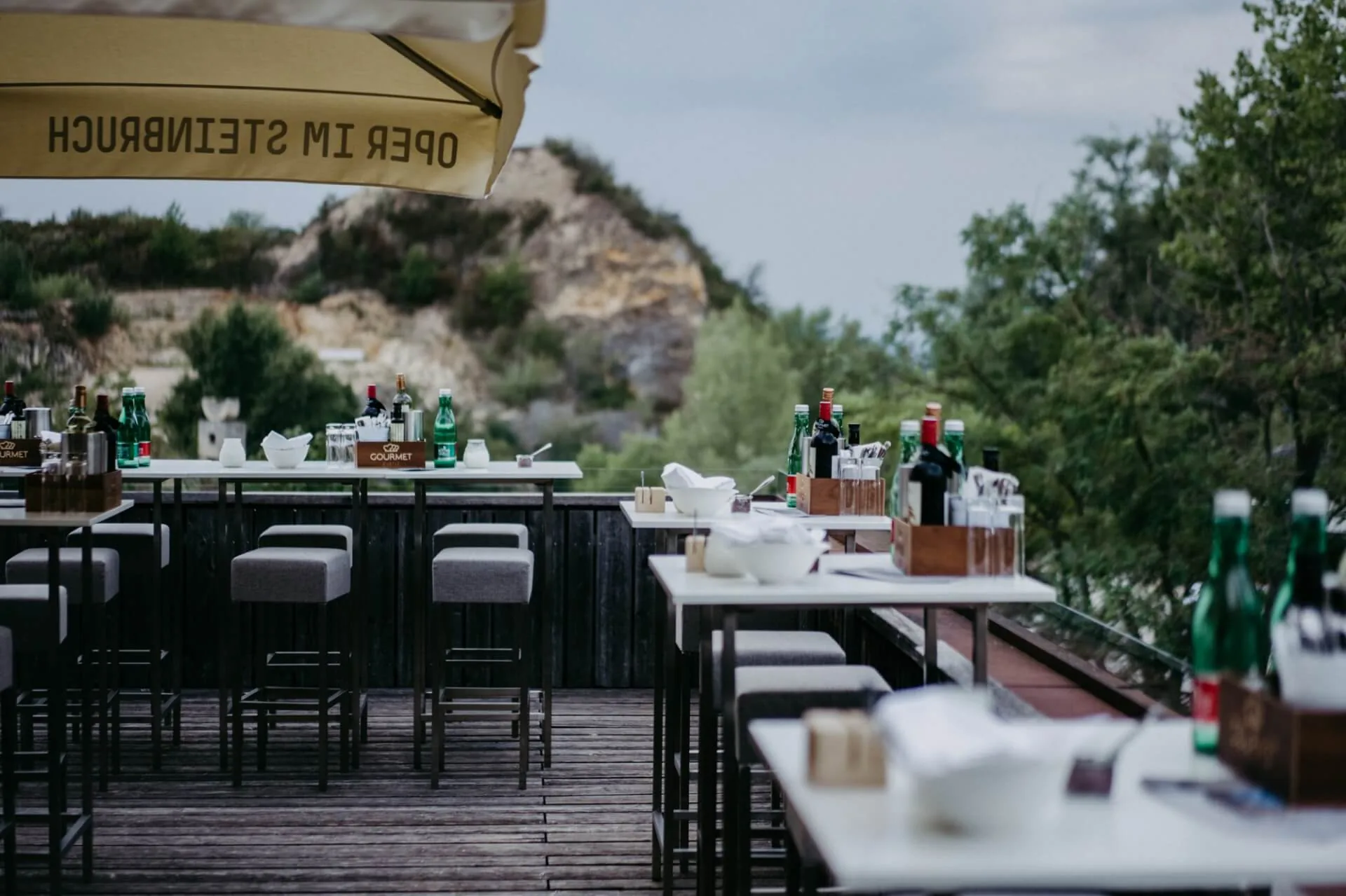 Restaurantterrasse im Freien mit Tischen und Hockern zum Essen, Wasser- und Weinflaschen auf den Tischen, umgeben von Grün und felsigen Hügeln im Hintergrund unter einem bewölkten Himmel.