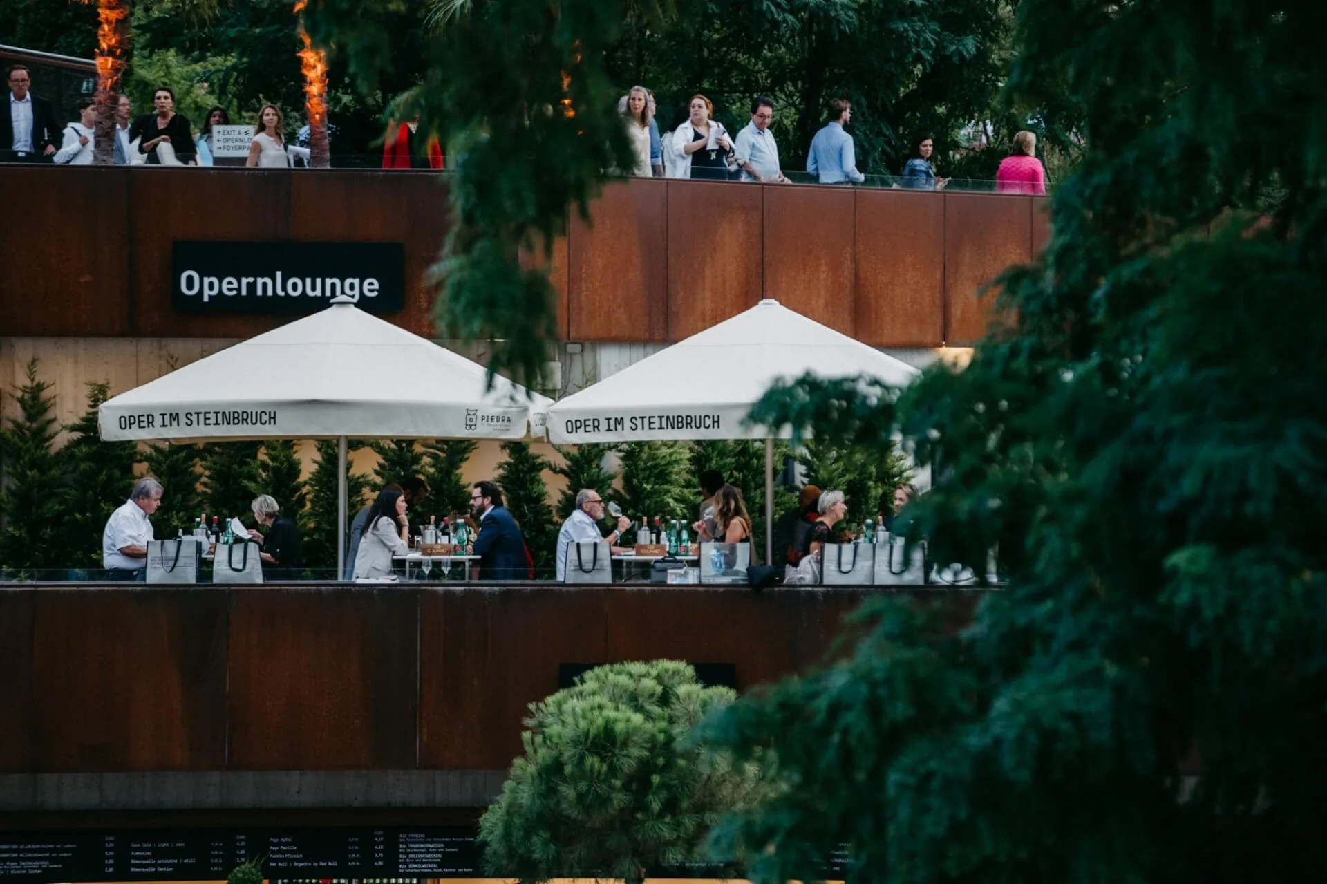 Auf zwei Außenterrassen der Opernlounge, die von Grünflächen umgeben ist, treffen sich die Menschen und unterhalten sich. Einige sitzen unter großen weißen Sonnenschirmen, während andere auf der oberen Ebene stehen oder gehen.