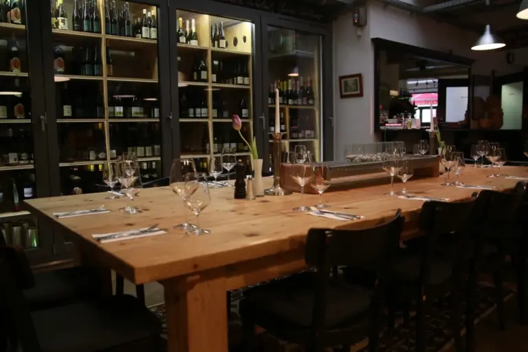 Magazin In einem gemütlichen Restaurant steht ein Esstisch aus Holz, auf dem Weingläser, Teller und Besteck stehen, umgeben von Stühlen. Im Hintergrund sind Weinflaschen in Regalen ausgestellt.