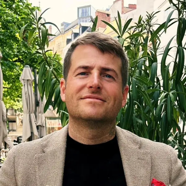 Ein Mann mit kurzen braunen Haaren und hellem Bart, der einen beigen Blazer und ein schwarzes Hemd trägt, sitzt im Freien in der Nähe von hohen Grünpflanzen mit Gebäuden und Regenschirmen im Hintergrund.