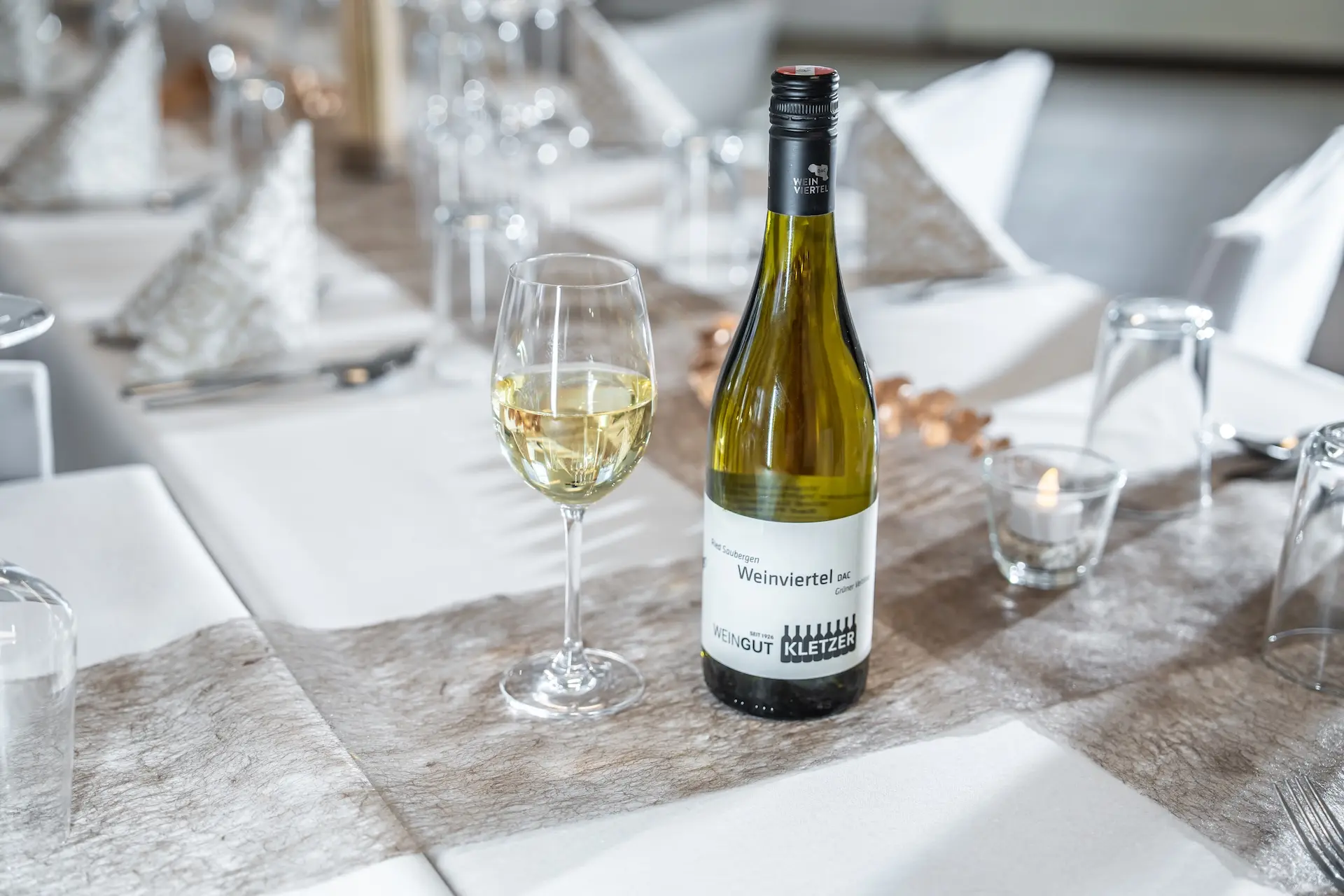 Eine Flasche Weißwein mit der Aufschrift Weinviertel und ein gefülltes Weinglas stehen auf einem eleganten, weiß gedeckten Esstisch mit gefalteten Servietten, Gläsern und einer kleinen brennenden Kerze im Hintergrund.
